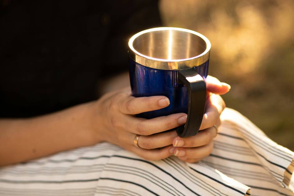 Hand holding a brushed stainless steel coffee mug against a backdrop of potted herbs and natural light